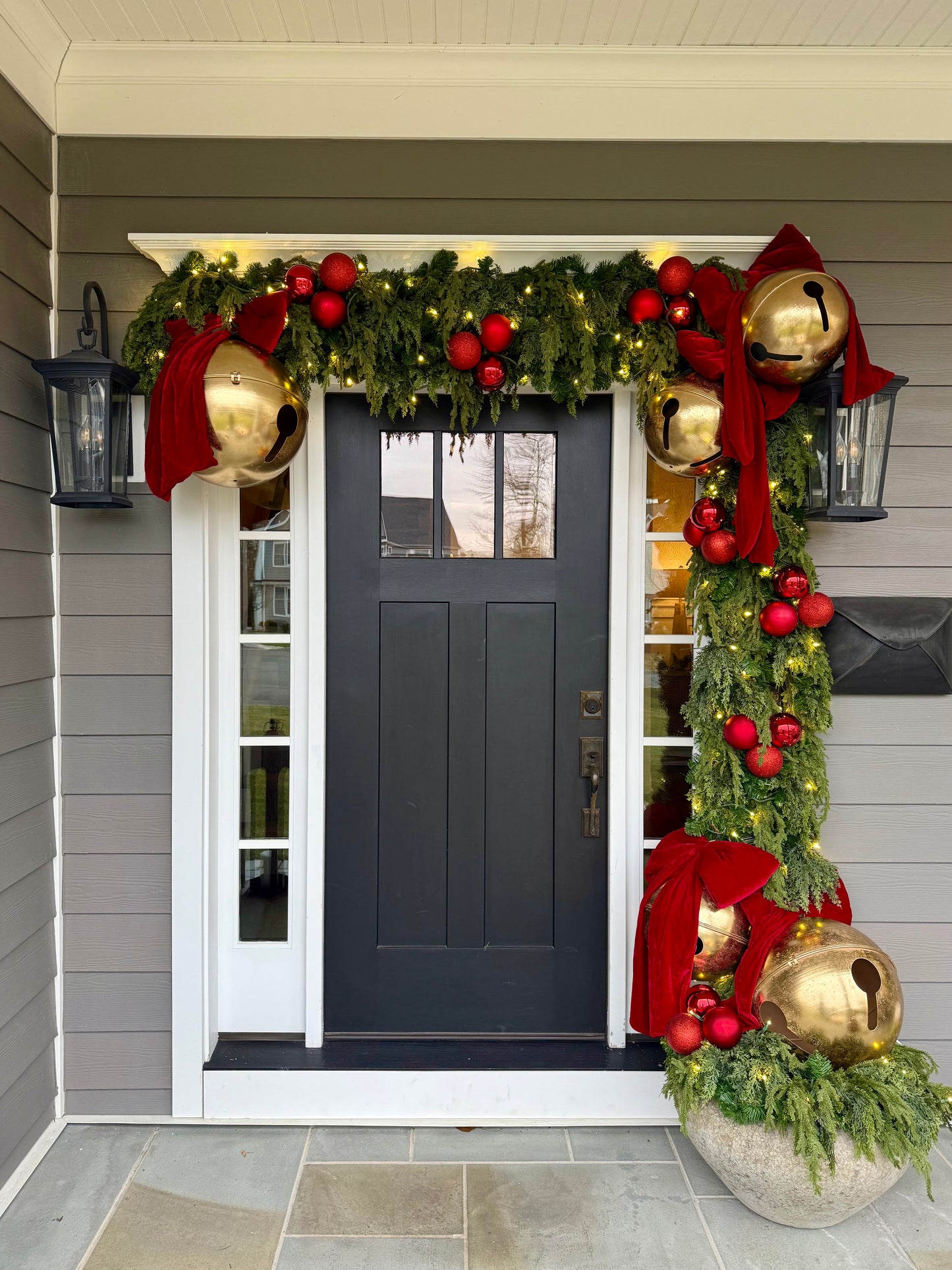 A festive Christmas front door decorated with green garlands, red ornaments, large gold bells, and red bows, set against a gray house exterior.