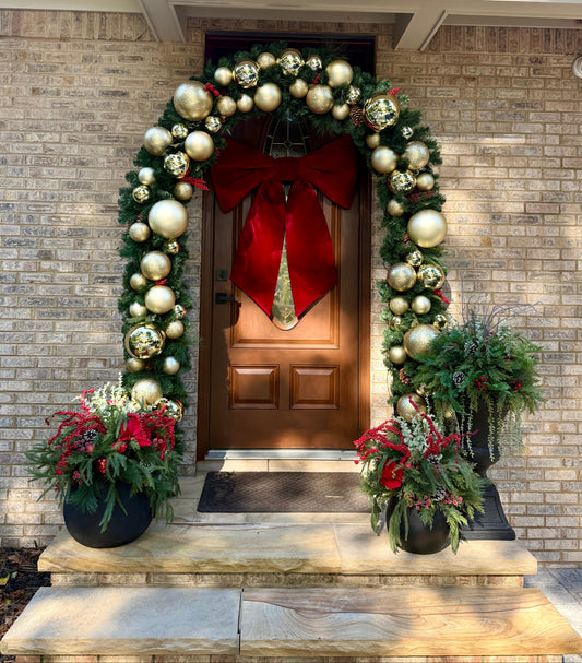 Decorative Christmas wreath with gold and silver ornaments and a red bow on a door, flanked by potted plants.